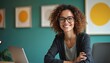 © Maryna - Confident woman with curly hair sits at desk in modern office. Dressed in casual attire, smiling, looking directly at camera. Gray sweater, white collared shirt, black pants, computer on desk.