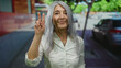 © Krakenimages.com - Senior woman with grey hair smiles in casual white shirt, standing outdoors on a street showing peace sign with an urban background of trees and cars.