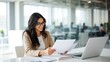© DESIGN MOSAI - Smiling young businesswoman wearing glasses reviews documents at her desk with a laptop in a modern office setting