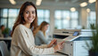 © miss irine - Young office worker with long brown hair sitting at desk in office setting. Wearing light gray sweater, smiling typing on white office computer. Neutral color scheme, window, plant in background.