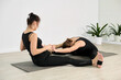© TRAVELARIUM - Two young women practice partner yoga in studio. They sit on mats, stretching forward in synchronized pose. Light walls and plants create calming ambiance