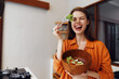 © SHOTPRIME STUDIO - Happy young woman in orange shirt enjoying fresh salad in a bowl, promoting healthy eating and vibrant living in a cozy kitchen setting