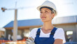 © Dreamera - Mujer joven con casco blanco y chaleco reflectivo, de pie en zona de construcción bajo el sol, expresión decidida y fondo urbano con estructuras metálicas