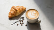 © HunterSnap - Minimalist flat lay of a fresh croissant and a latte with beautiful foam art on a light-textured stone background.