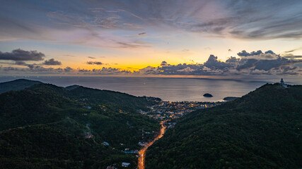 Wall Mural - Aerial hyper lapse view of A breathtaking sunset paints the sky in vivid hues of orange and blue over a coastal town, with lush green hills framing the scene. cloud in dramatic sunset over the ocean