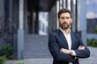 © Liubomir - A handsome businessman in a suit stands confidently with arms crossed in front of a modern office building.