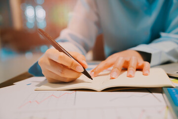 Poster - Hands Writing Notes with Pen in Notebook on Table Surrounded by Colorful Charts and Graphs in Modern Workspace