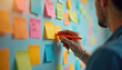 © Viktor - Business team uses Agile method for project planning. Man writes on sticky note, brainstorming ideas. Colorful sticky notes attached to wall. Agile, Scrum, Lean frameworks for teamwork.