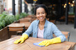 © Kowit - A smiling woman wearing yellow gloves wipes a wooden table in an outdoor cafe setting.