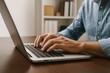 © Jony - Focused male hands typing on sleek silver laptop, wooden desk surface, bookshelf in background creating calm, productive workspace ambiance for efficient work.