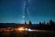 © anatoliy_gleb - Blurred silhouettes of tourists resting at campsite around campfire with glowing tents nearby under starry sky. Milky Way stretches above, casting magical light over tranquil mountain landscape.