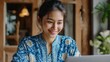 © sapiduduk - Smiling woman in batik browsing laptop in a warm modern workspace Image 4K UHD