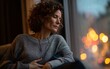 © Korkiat - A middle-aged woman with curly hair, wearing jeans and an oversized sweater, sits in the living room of her home at night, looking out through large windows.