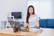 © Tj - Young Asian businesswoman working from home, seated at a desk while checking paperwork and using a laptop, with a stylish blue sofa creating a cozy backdrop in the living room