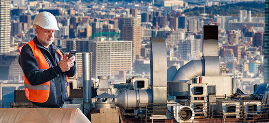 Poster - Man on roof with ventilation pipes. Worker servicing vents equipment. Man engineer with phone in front ventilation shaft pumps. Ventilation repair specialist in orange vest. Split, air conditioning