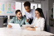 © fizkes - Three colleagues gathered around conference table, looking at tablet, hold document, reviewing information, discussing ideas while working on joint project together using digital and print resources