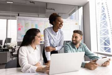 Group of three teammates engaged in positive communication in workplace using laptop and modern tablet device, laughing on joke during break gather in contemporary high-rise office building. Teamwork