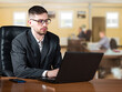 © Grispb - Businessman in suit working on laptop at office desk with colleagues in background, focused on corporate tasks and digital workflow. - Business. Office work. Management.