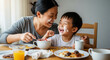 © SLCO LLC - Asian mom and child laughing with whipped cream on their faces at the dining table, breakfast mess in front of them, moment of shared silliness and joy.