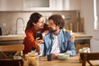 © Stockphotodirectors - A couple smiles at each other while looking at a smartphone in their cozy kitchen. They are seated at a wooden table enjoying breakfast in a warm and inviting atmosphere.