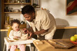 © pressmaster - Black young adult man feeding baby girl in high chair during daytime in modern kitchen, father preparing meal and interacting with child, concept of paternity leave and childcare