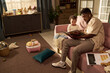 © pressmaster - Black young adult man sitting on floor reading storybook to toddler girl in living room during paternity leave, toys and documents scattered around, child listening attentively