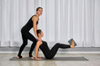 © TRAVELARIUM - Two women engaged in yoga session, one instructing with care. Set in light, airy studio, clad in black attire, creating calm, focused ambiance