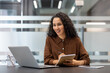 © Liubomir - A woman with curly hair smiles while taking notes during a video call on her laptop in an office setting.