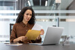 © Liubomir - A concerned businesswoman reviews documents while working on a laptop, suggesting work stress and business issues.