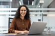 © Liubomir - A smiling woman with curly hair and glasses works on a laptop at her desk in an office setting.