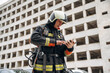 © standret - Tablet in hands. Professional firefighter is standing outdoors against big unfinished building