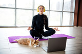 Young woman meditating on a yoga mat with her dog, headphones on, streaming guided meditation session on a laptop Young woman meditating on a yoga mat with her dog, headphones on, streaming guided meditation session on a laptop