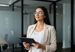 © shahida  - A confident young businesswoman wearing glasses and a beige blazer holds a tablet while working in a modern office environment