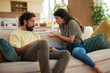 © Dexon Dee - Young couple having an argument while sitting on a sofa at home. The woman appears upset and is gesturing with her hands, while the man listens with a serious expression.