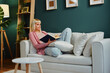 © djile - Young woman relaxing on sofa reading a book in cozy living room