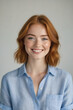 © f_bossa - A smiling young woman with auburn hair and fair skin gazes directly at the camera, wearing a light blue button-down shirt.