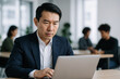 © Sandor - Serious asian businessman in formal suit working on laptop at modern office with diverse colleagues in background