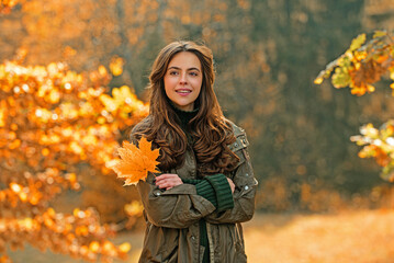 Wall Mural - Autumn woman with fall yellow maple leaf near face, outdoor portrait. Girl with autumn leaves. Portrait of beautiful young woman outdoors in autumn park. Autumn mood. Autumnal fall concept.