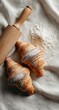 © Selsi - Two golden croissants dusted with powdered sugar rest on a linen cloth alongside a wooden rolling pin and scattered flour (1)