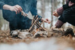 © qunica.com - A close-up photograph depicting two individuals collaborating to construct a campfire amidst an outdoor natural setting, conveying themes of wilderness, survival, and companionship.