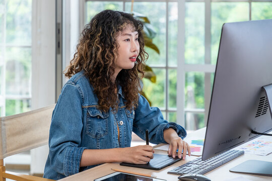 Asian female graphic designer using drawing tablet and desktop in creative workspace