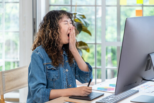 Tired Asian graphic designer yawning at desk with drawing tablet and computer