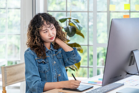 Asian female graphic designer with neck pain working at computer with digital tablet, office syndrome