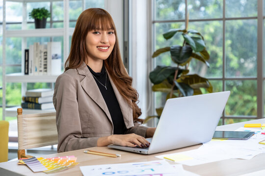 Young Asian woman smiling while working on laptop with design documents and color swatches on creative desk in modern office, looking at camera