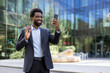 © Liubomir - An African American businessman waves while video calling on his smartphone outside a modern office building, smiling and greeting someone on the call.