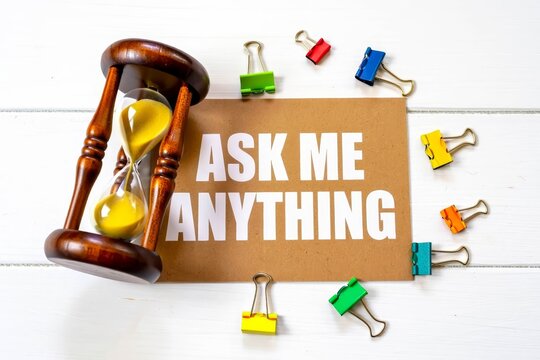 Ask Me Anything text on paper card with hourglass and stationary on wooden background.