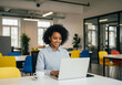 © Peeradontax - A happy young Black businesswoman with curly hair is seated at a desk in a modern, brightly lit open-plan office.