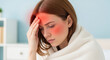 © SABBIR - Woman wrapped in light blanket touching forehead with red glow and flushed cheeks symbolizing high fever or chills full head and upper torso in soft blurred background