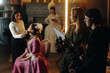 © pressmaster - Caucasian young adult woman sitting in chair receiving makeup from another woman while film crew preparing camera on Baroque Renaissance set