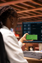 Poster - Woman using mockup smartphone and AI LLM chatbot on computer screen in home office. African american person holding chroma key phone uses artificial intelligence chatbot on monitor